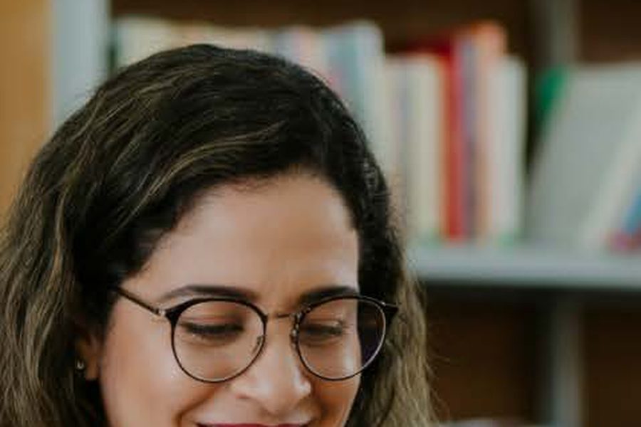 Professora sorrindo enquanto acompanha as respostas da turma pelo celular, com o caderno aberto sobre a mesa.