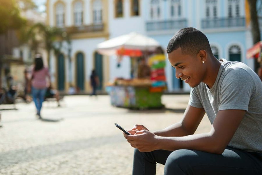 Jovem sentado na rua de um centro histórico, estudando pelo celular.